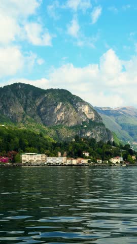 Elegant historical hotels and buildings along the water in Griante village at Lake Como with majestic green mountains and blue sky in background.