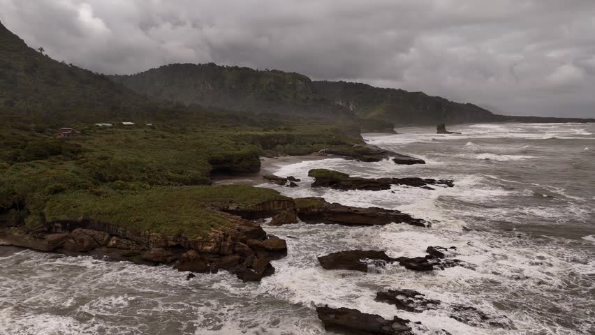 Dramatic and wild rocky coastline with waves crashing on moody, overcast day on Truman Track, New Zealand. Aerial drone panoramic view