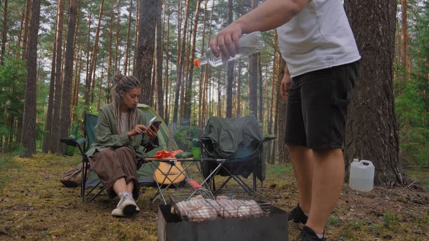 Relaxed young couple enjoying a picnic in the woods while the bearded man grills meat on a charcoal barbecue and the woman with dreadlocks sits nearby using her smartphone in the background