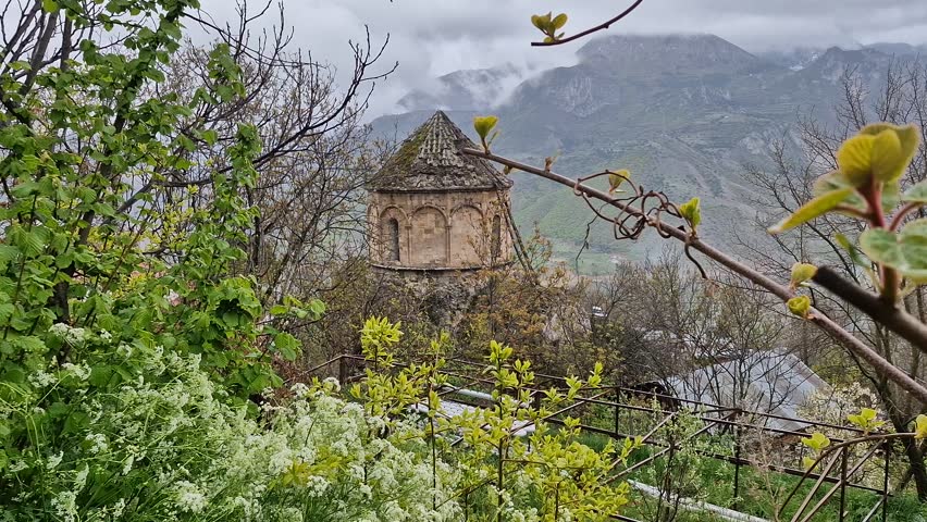 Stone medieval chapel standing on a hill surrounded by lush green trees with a mountain background