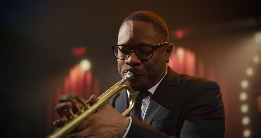 Portrait Black Male Trumpeter in Glasses Leads Live Jazz Band Concert at a Bar, Playing a Powerful Song on Stage as the Audience Enjoys the Music Event and Follows His Dynamic Solo Performance.