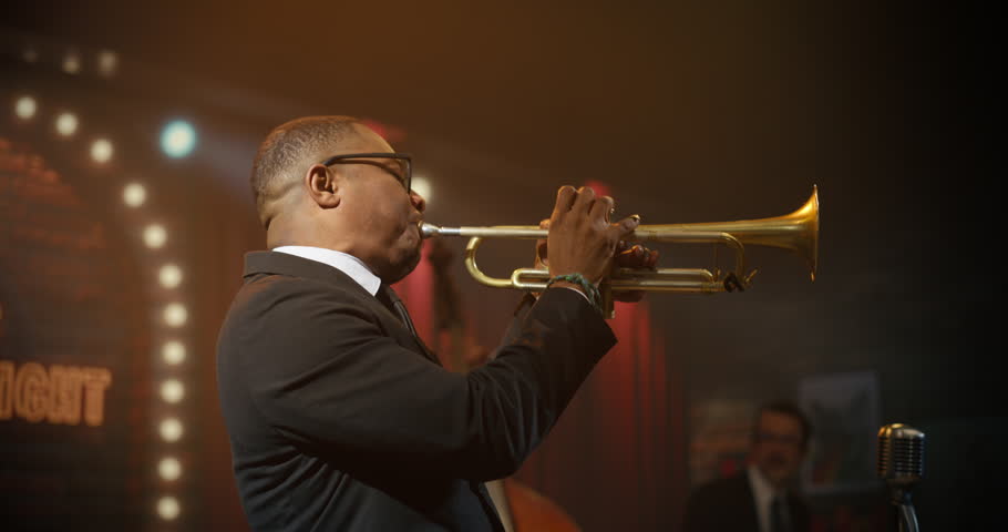 Black Male Trumpeter in Glasses Performs a Soulful Jazz Song Live With His Band on a Bar Stage, Playing an Energetic Concert Set as the Audience Enjoys the Intimate Music Event.