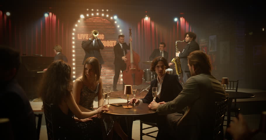 Four Young Adult Friends Relax at a Small Table in a Live Jazz Bar, Chatting, Smiling and Raising Drinks While the Band Plays Nearby, Fun Night of Music, Friendship and Connection.