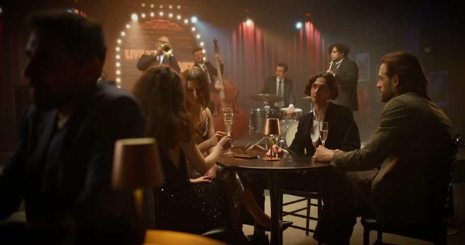 Four Young Adult Friends Enjoy a Live Jazz Bar Night, Sitting at a Small Table, Sipping Drinks, Talking, Smiling and Sharing a Relaxed Social Evening While the Band Performs on Stage Nearby.