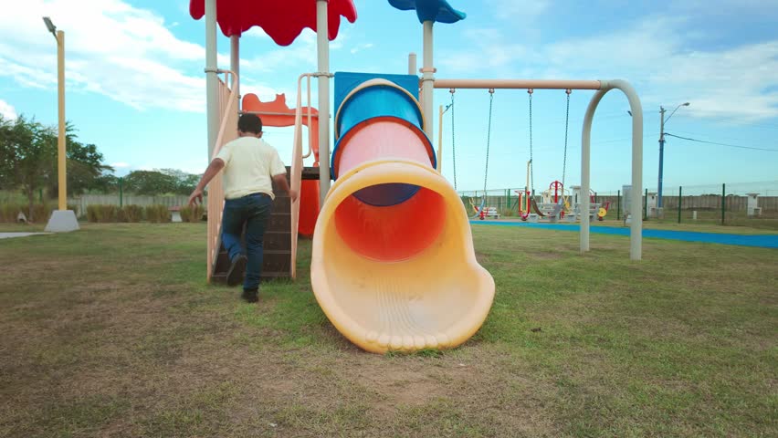 Cheerful elementary school-aged boy happily sliding down a colorful plastic tube slide at an outdoor park, enjoying a beautiful sunny day