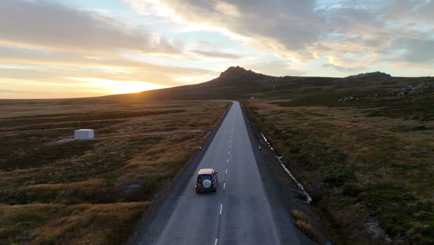 Cinematic aerial view of a car driving through remote coastal landscape at sunset in the Falkland Islands, tracking drone footage. Into the sun.