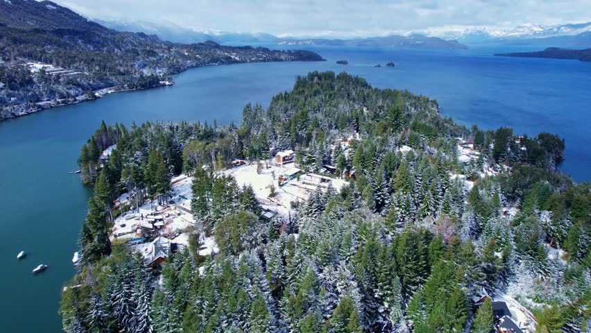 Static drone shot with a vertical tilt up camera movement, starting from snow-covered pine trees to reveal the blue Nahuel Huapi Lake and snowy mountains in Puerto Manzano, Patagonia.