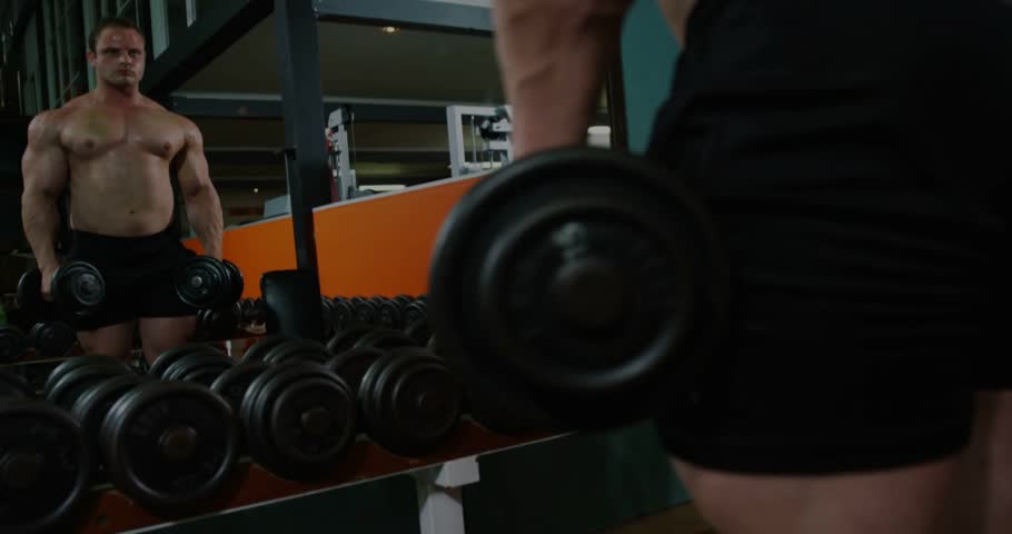 Checking form shirtless male in black shorts doing lateral dumbbell raises at gym near mirror,rack. Shoulders, muscular, strength, training, fitness, back, arms