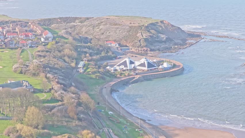 North Bay in Scarborough North Yorkshire England shows SEA LIFE Scarborough pyramidal buildings on coastal promontory with beach, headland and North Sea shoreline, aerial drone forward flight shot