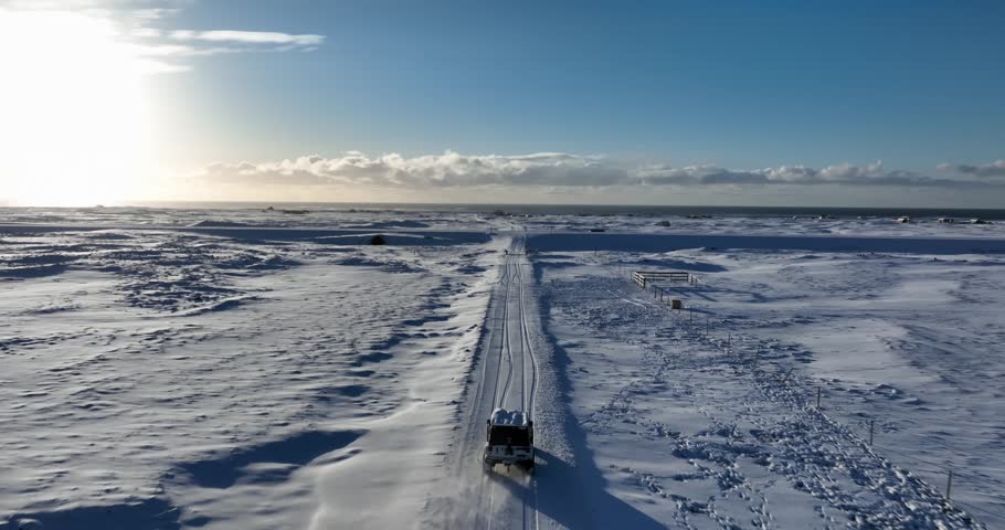 Aerial view of a lone vehicle driving along a snow-covered road across the Reykjanes Peninsula, Iceland, with vast winter plains and bright low sun on the horizon.