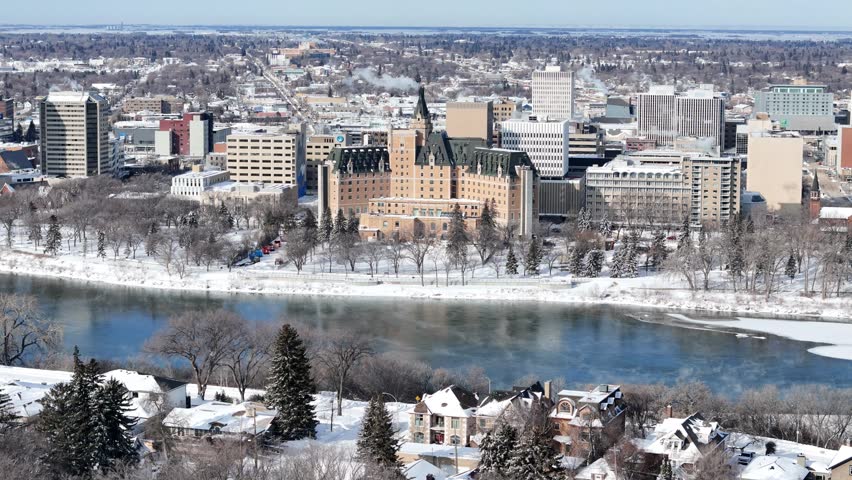 Aerial drone footage captures downtown Saskatoon in winter. Snow blankets streets and river banks. Historic and modern buildings line the river