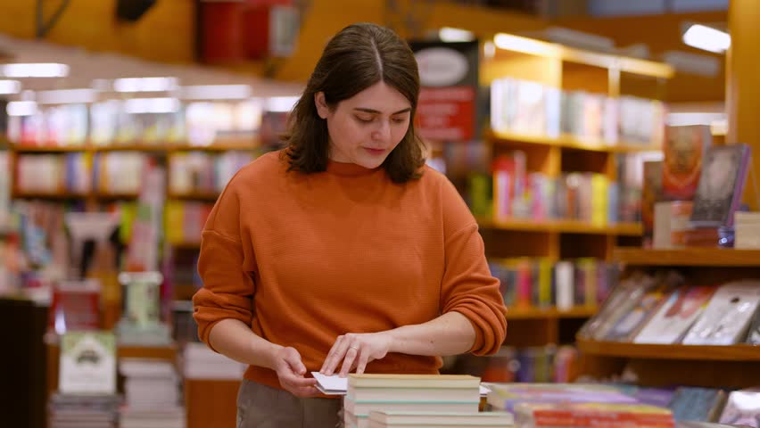 Latin Woman in Orange Top Browsing and Reading Books on Display Table in Bookstore Retail Store Medium Shot