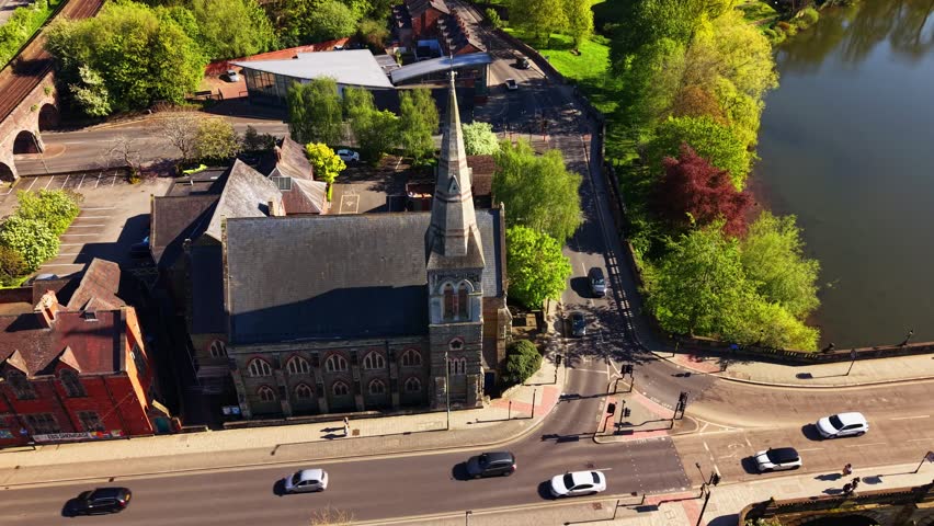 Drone orbiting Victorian Gothic church with tall spire at busy crossroads in Shrewsbury town centre. Railway viaduct, red brick buildings and traffic visible below.