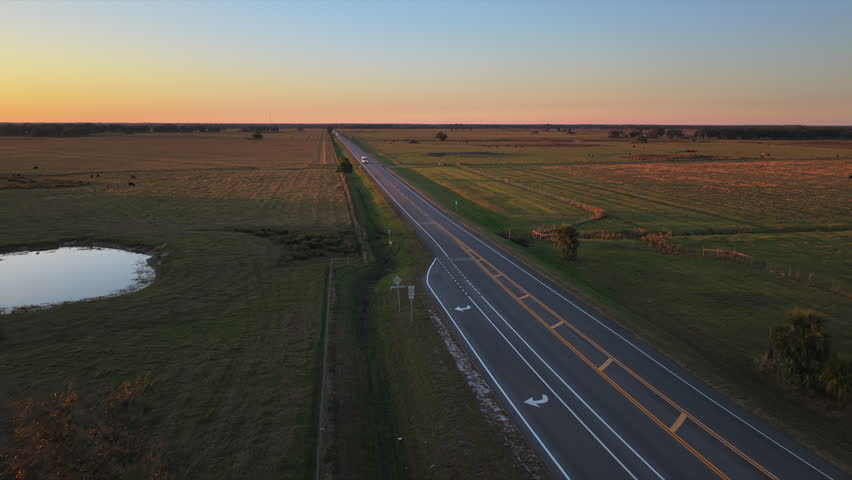 Aerial view of rural highway at sunset with cars driving fast along a straight asphalt road with countryside landscape.