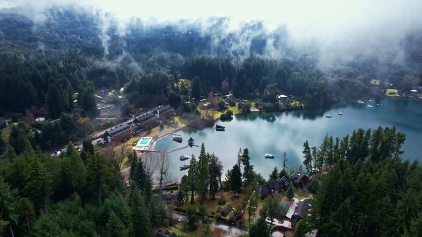 Drone moving forward over a perfectly reflective Nahuel Huapi Lake, approaching a luxury mountain resort as swirling low-lying clouds and mist emerge from the surrounding dense pine forests in Patagon