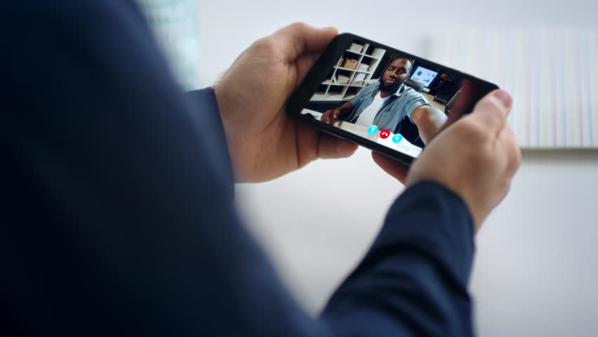 Man hands video calling smartphone at office closeup. Unrecognizable manager holding mobile phone talking with african american smiling colleague. Unknown guy looking partner screen greeting online