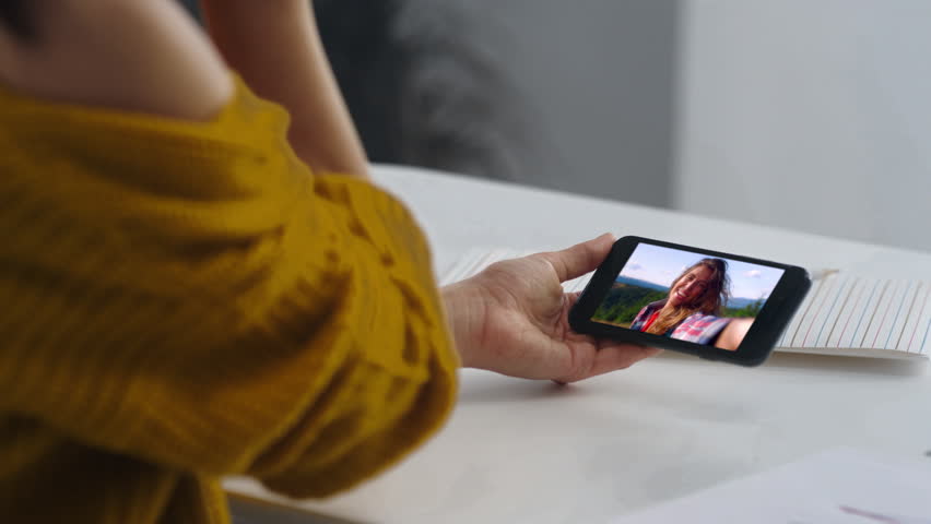 Woman watching smartphone screen at white table closeup. Young lady hands holding mobile phone enjoying travel blog video at home kitchen. Relaxed brunette face involved in online conference alone