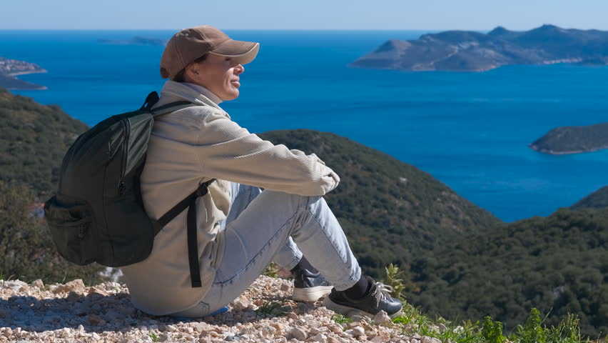 Hiker woman resting on a mountain enjoying the sea view. Smiling woman sitting on a mountain top with a backpack, relaxing and enjoying the breathtaking view of the sea and islands on a sunny day