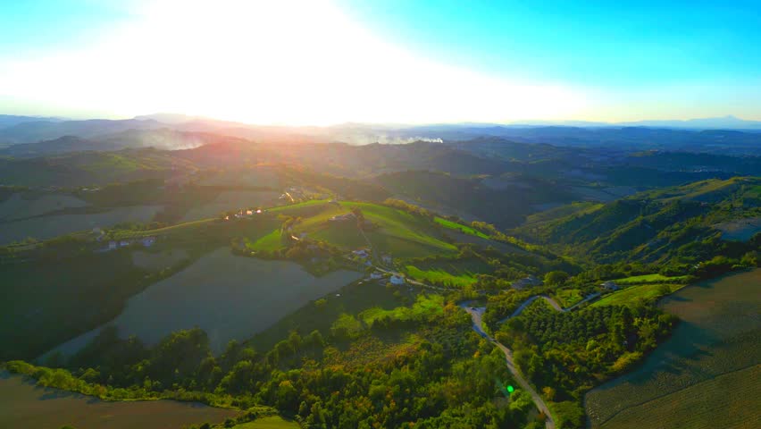Acrobatic aerial pullback pan left truck left shot from Monterubbiano, the hills surrounding the town centre in full display, with gentle carpety lawns, thickets and roads, mountains in the distance