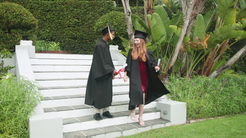 Graduation: two grads stepping on lawn with diplomas, man popping popper, overlays around them. Alumni, celebration, confetti, garden, tropical, hedge, steps