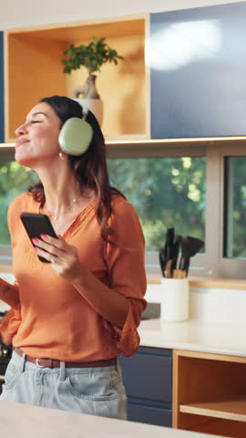 Happy woman, headphones and dancing with phone in kitchen with sound, streaming and song in home. Person, smile and rhythm with mobile app, audio and listen to music with subscription at apartment