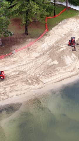 Aerial view of a red excavator moving sand for beach restoration along a waterfront canal