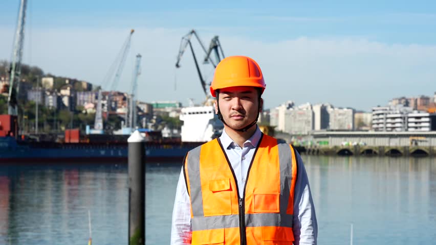 Asian male engineer in a hard hat and safety vest standing with arms crossed at a commercial harbor, looking at the camera with a confident smile against a background of cranes and cargo ships