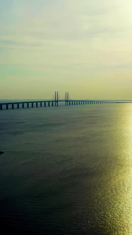 Vertical video. Oresund bridge. The longest combined road and rail bridge in Europe spans the Oresund strait, connecting Denmark and Sweden in a single sweeping structure. The video captures the bridge from an elevated aerial perspective, showing the full length of the roadway and its twin cable-stayed pylons rising above the water. The scene is set during the golden hour, with warm sunset light creating a bright reflective path across the calm sea surface. The bridge extends from the Swedish coast toward the horizon, where it meets the artificial island of Peberholm before continuing as an underwater tunnel toward Copenhagen. The cable-stayed main span provides vertical clearance for maritime traffic, while the lower deck carries railway lines beneath the four-lane motorway above. The surrounding waters are tranquil, with soft gradients of blue and gold dominating the color palette. The composition emphasizes the engineering scale and linear geometry of the structure against the vast expanse of the strait. This landmark serves as a vital transport artery and a symbol of cross-border cooperation between the two Scandinavian nations. Aerial View. Rich colors