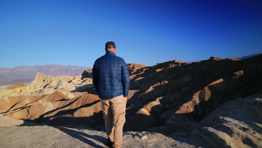 A man wearing blue puffer jacket and khaki pants stands with his back to camera overlooking stunning golden and brown eroded rock formations of Zabriskie Point in Death Valley National Park under.