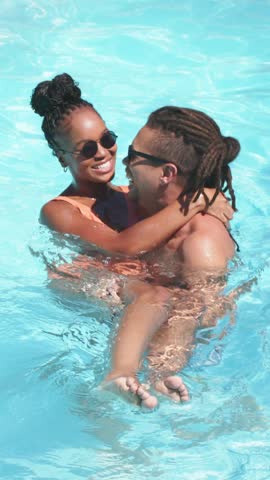 Vertical video: Lifting man cradling woman in outdoor pool for joy, wearing shades, orange swimsuit. Couple, romance, affection, water, sunlight, sunglasses, vertical video
