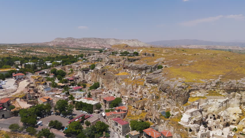 Urgup, Nevsehir, Turkey. A wide aerial perspective captures the dramatic convergence of ancient rock-cut architecture and modern settlement along the edge of a steep cliff face in the Cappadocia region. The scene reveals numerous cave dwellings and boutique cave hotels carved directly into the soft volcanic tuff, their arched openings and terraced facades blending into the pale, eroded rock. Below the cliff line, a compact town spreads across the arid plateau, with buildings featuring terracotta and ochre rooftops clustered among scattered green trees. The surrounding landscape is defined by rolling, golden-brown hills and distant mountain ridges under a bright, cloud-dotted sky, indicating a dry summer day. The camera maintains a steady, elevated vantage point that emphasizes the scale of the geological formations and the density of the historic settlement. This area lies within the Goreme National Park and Rock Sites of Cappadocia UNESCO World Heritage site, recognized for its vast troglodyte complexes where early Christians carved sanctuaries and villages into the rock to escape persecution, a tradition that has evolved into the region