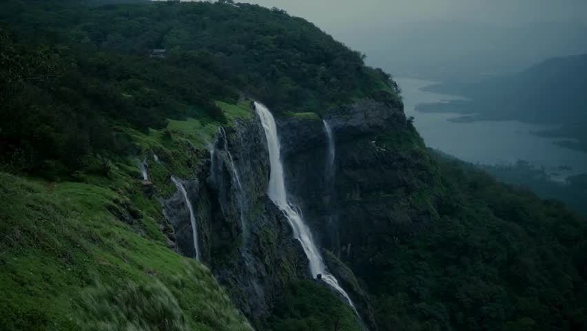 Breathtaking waterfalls flow dramatically from steep cliffs, surrounded by vibrant green vegetation and a tranquil lake view from above, creating a stunning natural landscape.