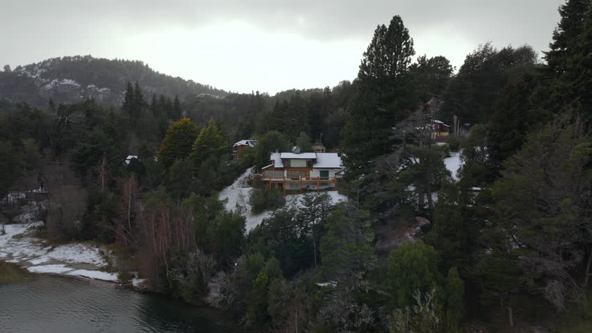 A solitary house sits atop a snowy hill surrounded by dense forest near a tranquil lake in Patagonia. Perfect for showcasing remote living or winter landscapes.