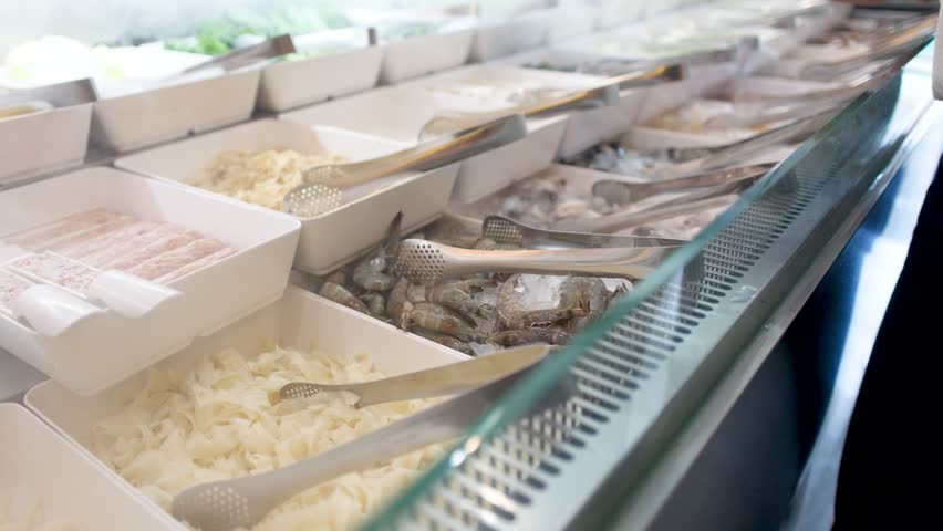 A person uses metal tongs to pick raw shrimp from an iced buffet display counter