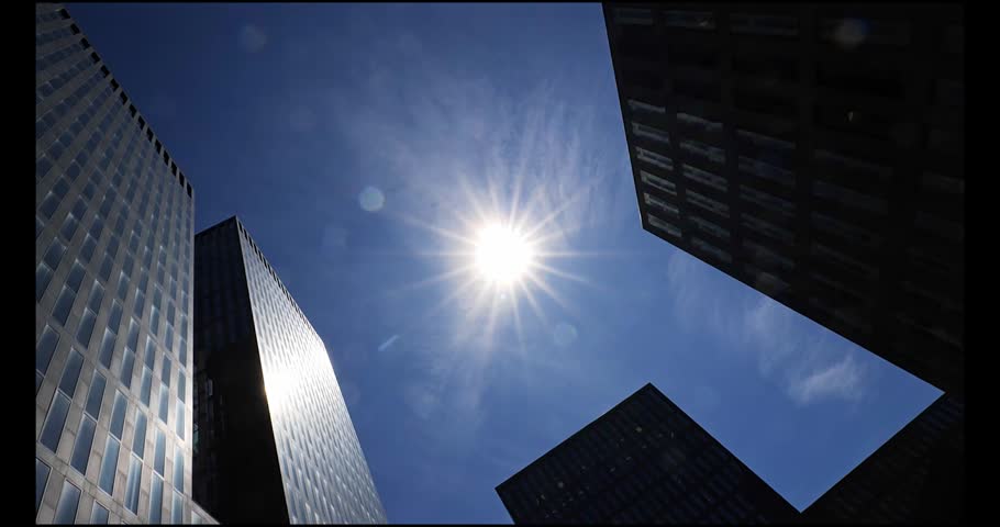 Cinematic low angle barrel roll shot looking up at a brilliant sunburst and blue sky between modern glass buildings in a city center