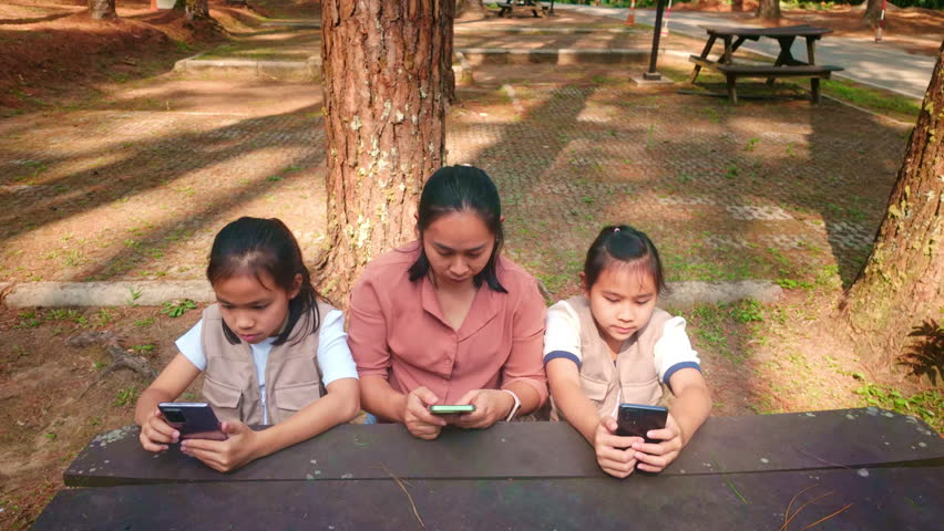 Asian mother and her young daughters sitting together in a pine forest using smartphones, family digital lifestyle and technology addiction concept.