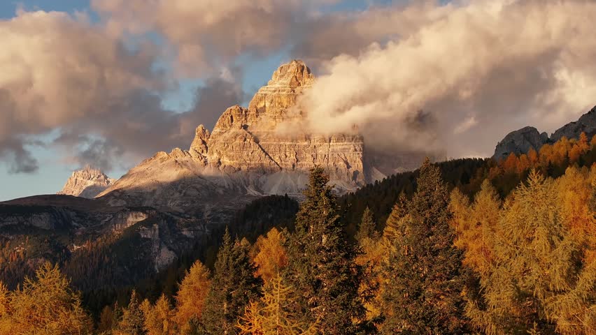 Drone view of Tre Cime di Lavaredo in the Dolomites at sunset. Iconic rocky peaks bathed in warm alpenglow with soft pink clouds above autumn forest in northern Italy.