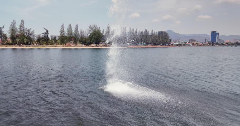 Large plume of water spraying from the Seahorse statue into the river in Kampot with a tree lined shore and distant mountains in the background.