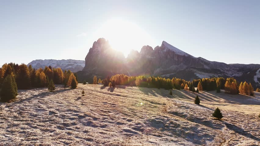 Stunning drone view of the Sassolungo mountain range, Langkofel in the Dolomites. Majestic alpine peaks covered in light snow, autumn colored forest all around, and yellow grass covered in morning white hoarfrost. Picturesque natural landscape