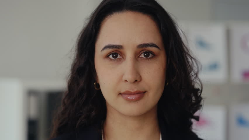 Closeup portrait of Hispanic businesswoman with dark loose hair stands in corporate office. Young female entrepreneur with calm expression in workplace