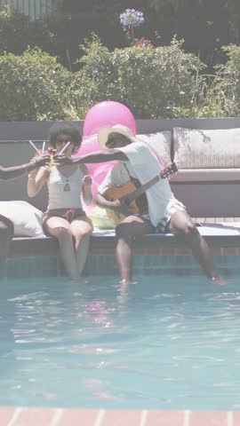 Vertical video: Guitarist reaching to clink, toasting at pool, tech overlay drifting across pair. Friends, outdoor, backyard, sunny, casual, acoustic, vertical video