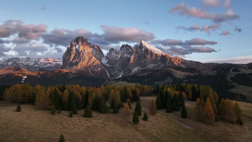 A stunning drone view of the Sassolungo, Langkofel mountain group in the Dolomites. Majestic alpine peaks covered with light snow glow in the warm evening sunlight, surrounded by autumn forest and green alpine meadows. Scenic natural landscape during golden hour in the Italian Alps.