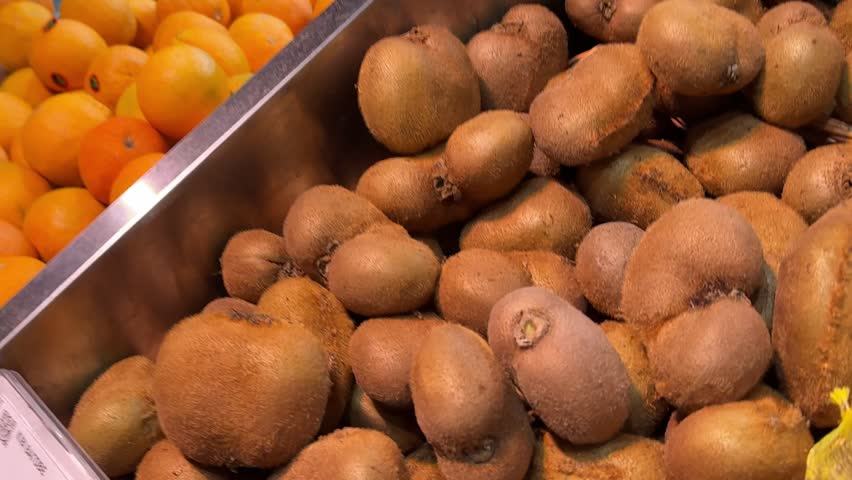 A large group of kiwi fruits are piled in a black crate at a market during the afternoon. They have a brown, fuzzy skin and various shapes.