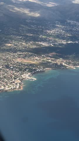 Aerial view of a coastal town with turquoise sea, dense housing, and mountain landscape, showing a peaceful Mediterranean shoreline from above.