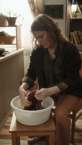 Vertical shot of woman washing clothes by hand, using basin, sitting on chair, cleaning garments in poor apartment