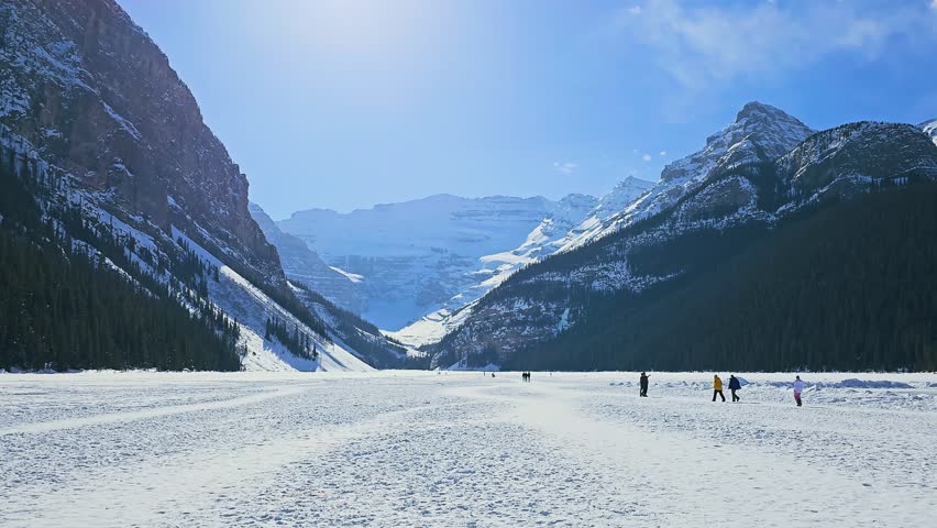 Expansive frozen Lake Louise in Banff, Alberta offers a peaceful winter scene with snow-covered terrain, distant people, dark forest, bright sky, and towering alpine mountains.