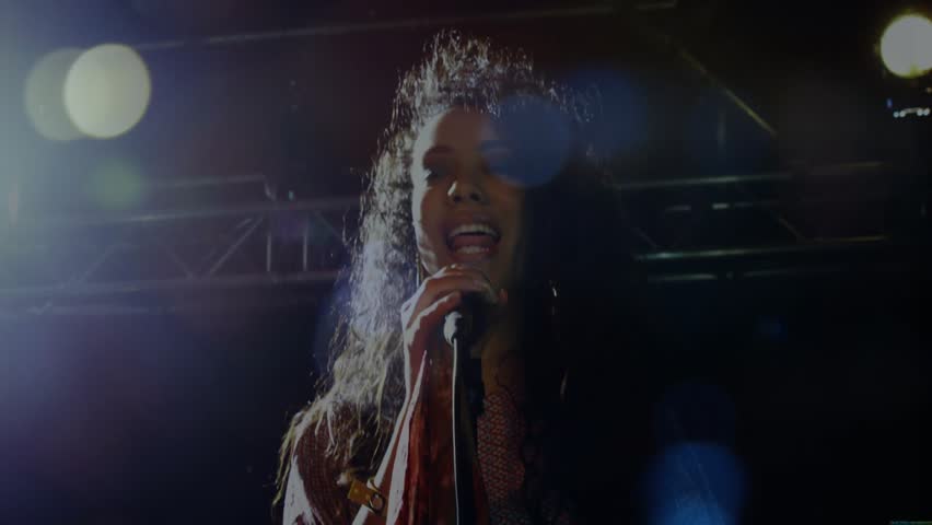 Singer starting singing into mic under lights, whipping hair, lens flares, energizing concert fans. Vocalist, microphone, energetic, onstage, motion, lighting, truss