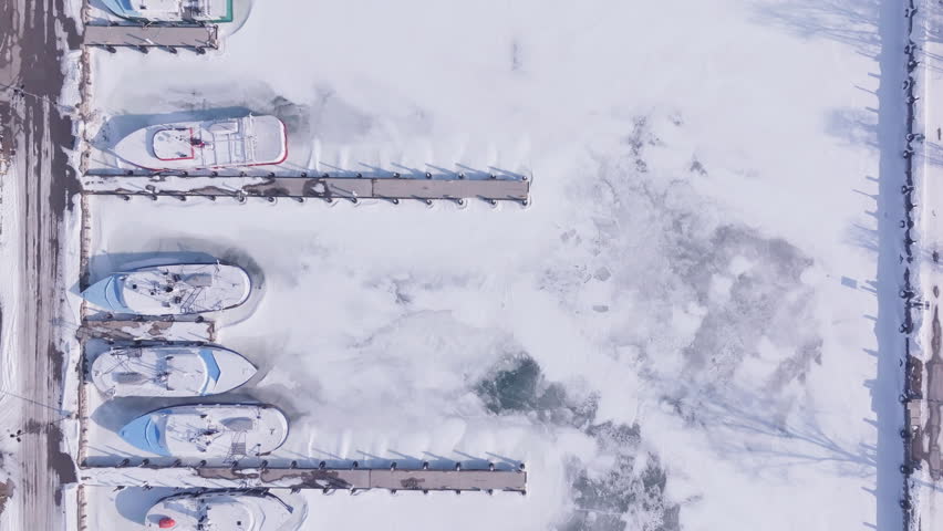 High-angle aerial view looking straight down at multiple commercial fishing boats in their slips. The marina is locked in ice and covered in snow. Port Dover, Ontario.