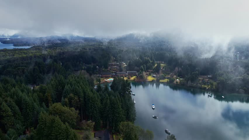 Forward drone dolly in shot over a tourist resort in Puerto Manzano, Villa La Angostura. Low clouds and mist covering the pine forest and Nahuel Huapi Lake in Patagonia Argentina.