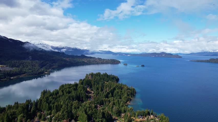 Forward drone dolly in shot showing the lush pine forests of a peninsula, surrounded by the deep blue waters of Nahuel Huapi Lake and snow-capped mountains under a cloudy sky.